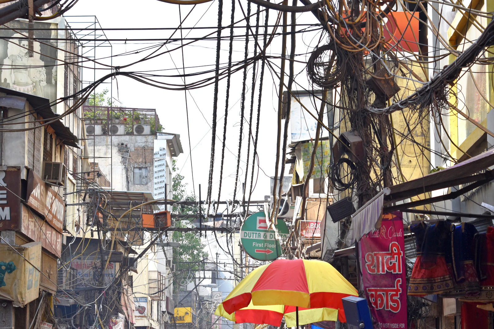 A crowded Old Delhi bazaar lane under a tangle of overhead power cables, with shopfronts and a red umbrella stall